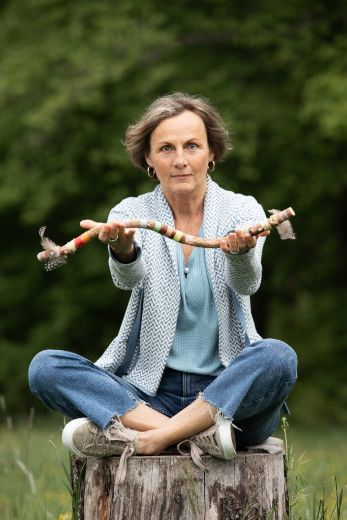 Petra Irana Persy auf einem Baumstumpf in der Natur mit schamanischem Redestab.