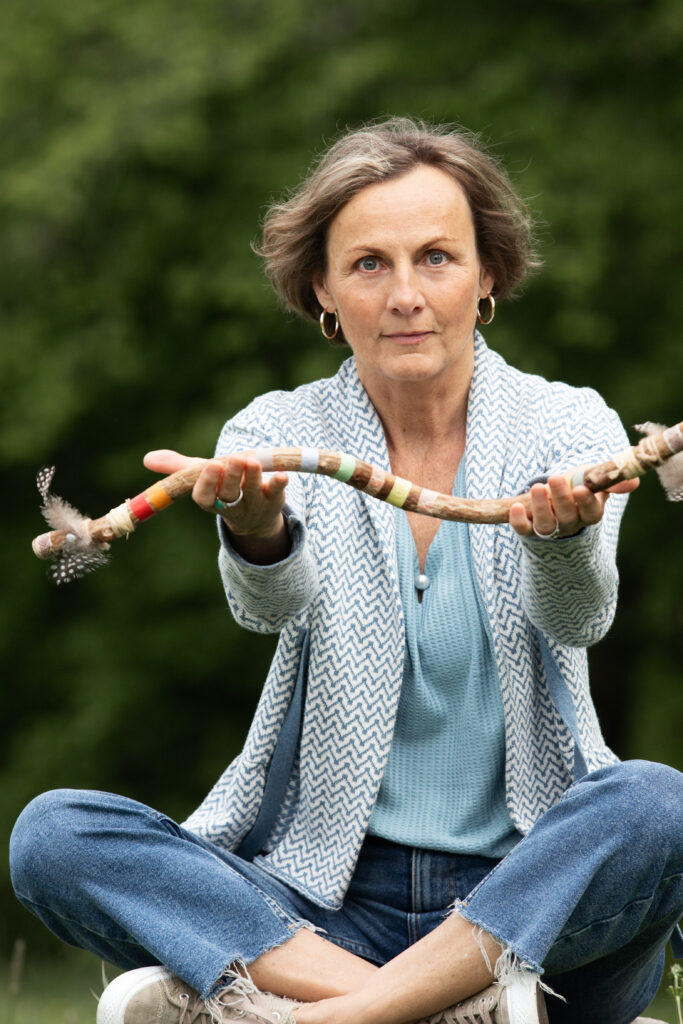 Petra Irana Persy auf einem Baumstumpf in der Natur mit schamanischem Redestab.
